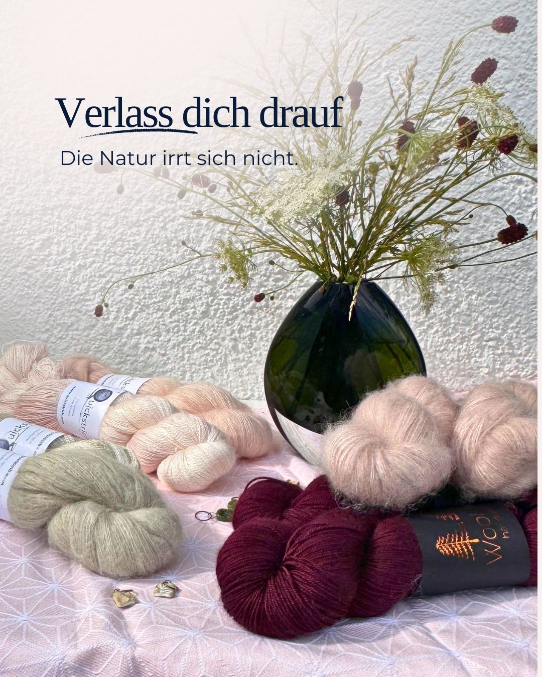 edle Wollstränge in den Farben Nude, Rose, grau und Weinrot, hübsch drapiert. Dahinter ein Strauß Wildblumen in einer grünen Vase.